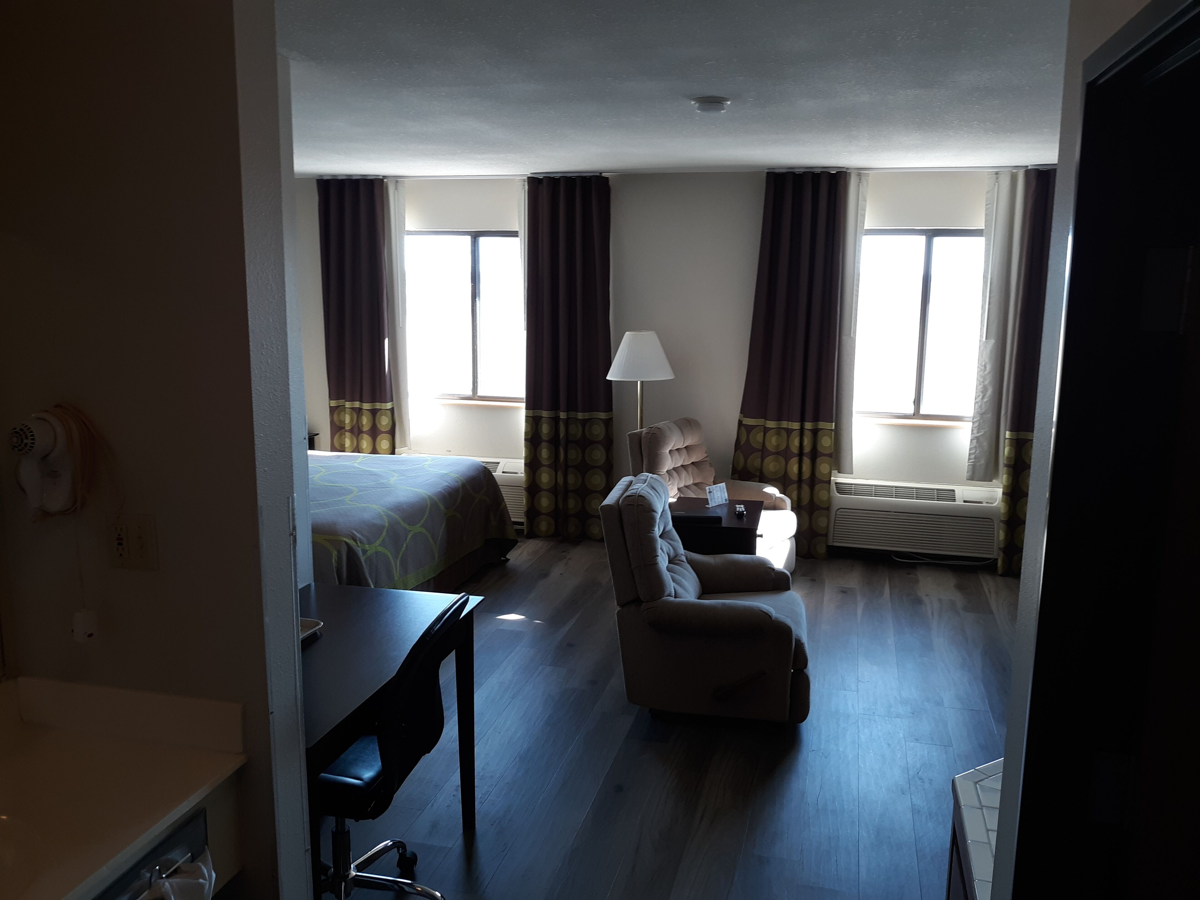 Una habitación de hotel con cama, escritorio, silla y una mesita. Hay dos ventanas con cortinas, una lámpara sobre la mesita y un equipo de aire acondicionado.