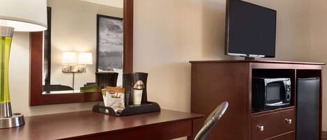 A hotel room with a wooden desk, a flat-screen TV, a microwave, a refrigerator, a chair, a lamp, and a framed picture on the wall.