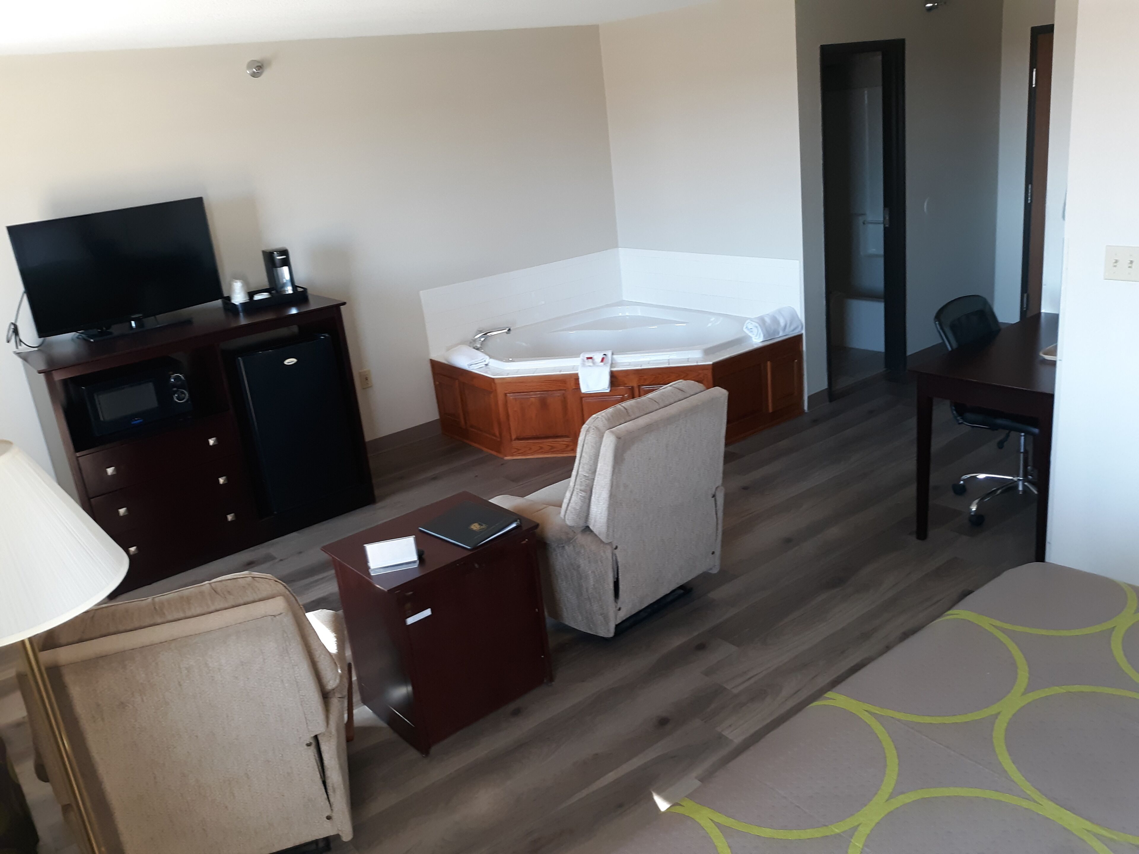 A hotel room with a TV, a minibar, a sofa, a desk, a chair, and a large bathtub.