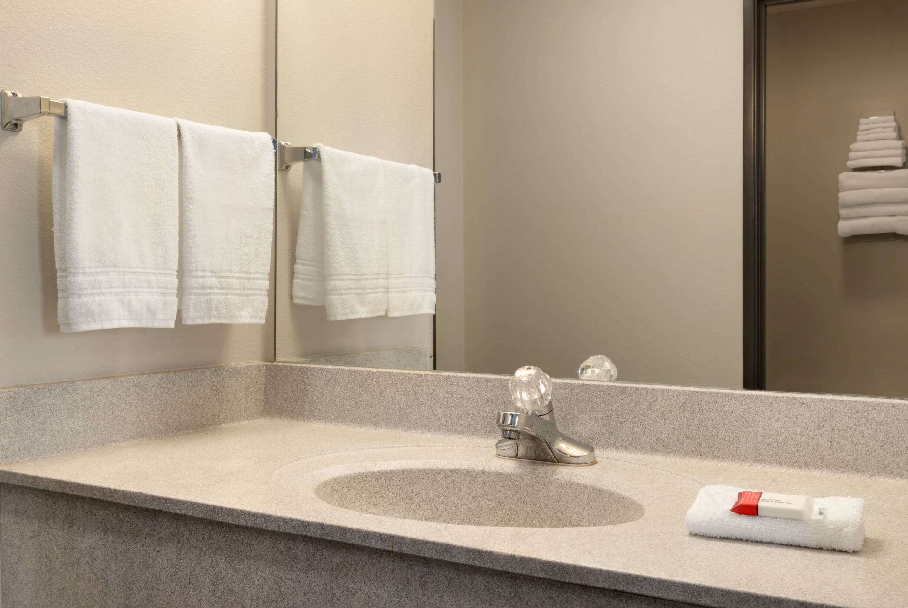 A bathroom with a granite countertop, a sink, and a mirror. There are white towels hanging on a rack and a folded towel on the countertop.