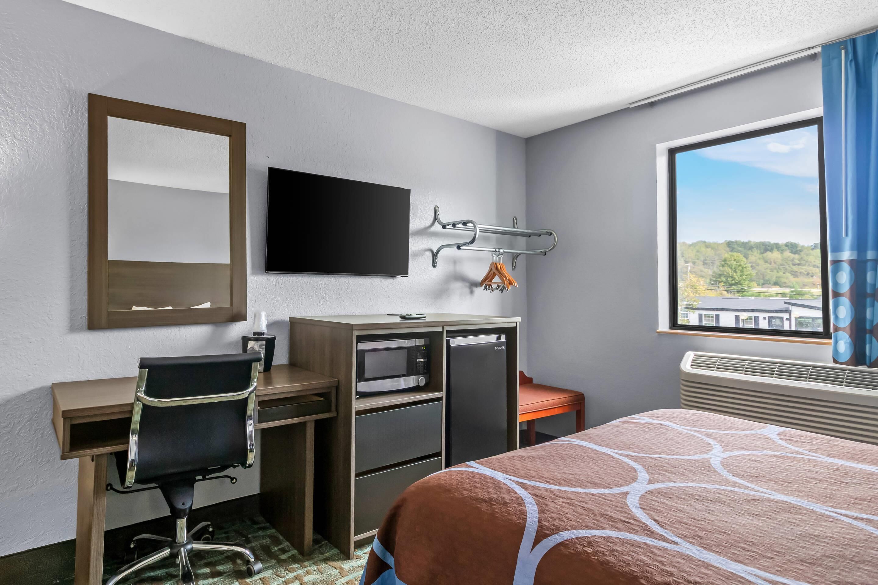 Habitación de hotel con cama, escritorio con silla, televisión, microondas, refrigerador pequeño y ventana con vistas a árboles y edificios.