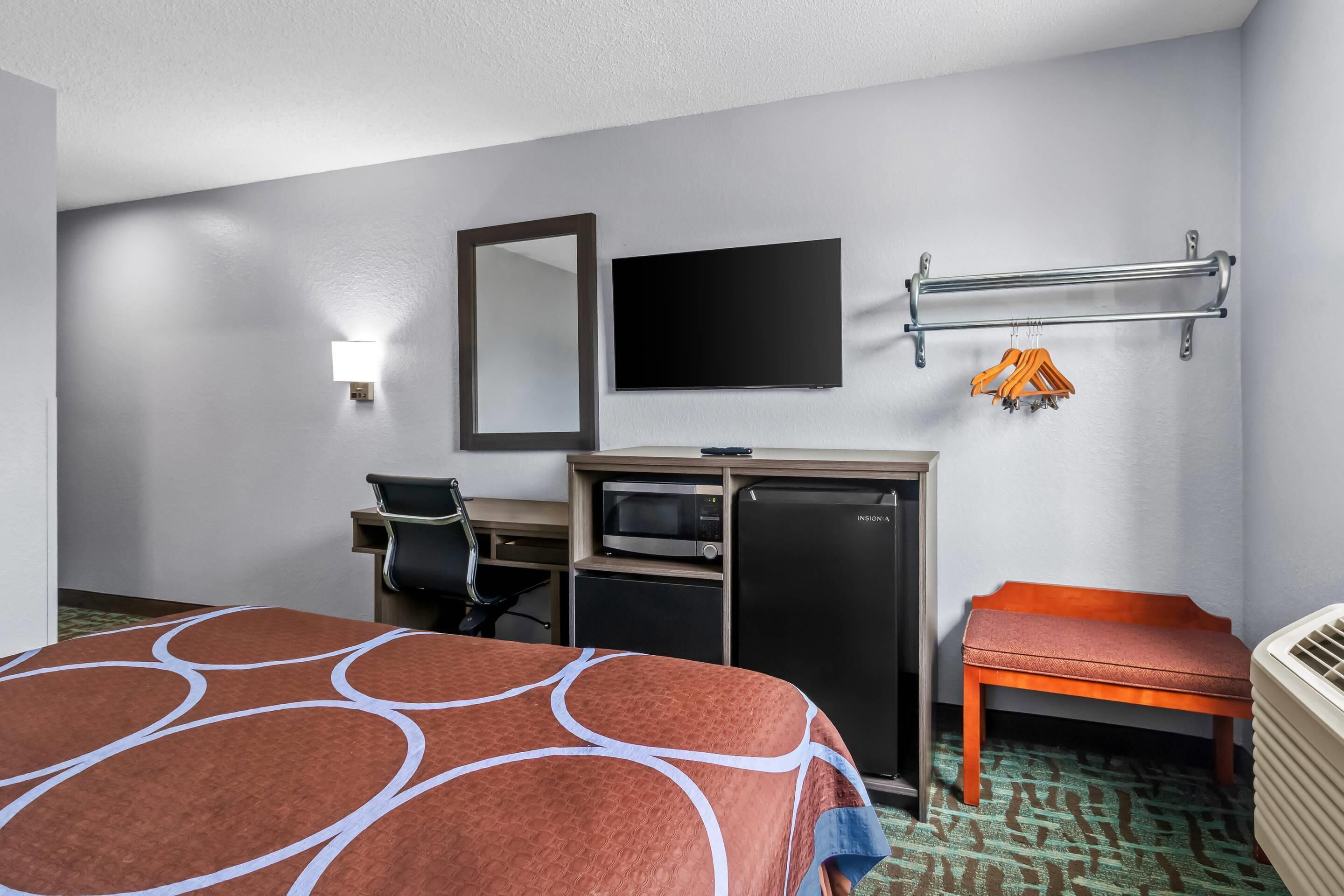 Quarto de hotel com cama, escrivaninha com computador, micro-ondas, frigorífico, televisão e um banco.
