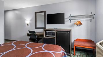 Quarto de hotel com cama, escrivaninha com computador, micro-ondas, frigorífico, televisão e um banco.