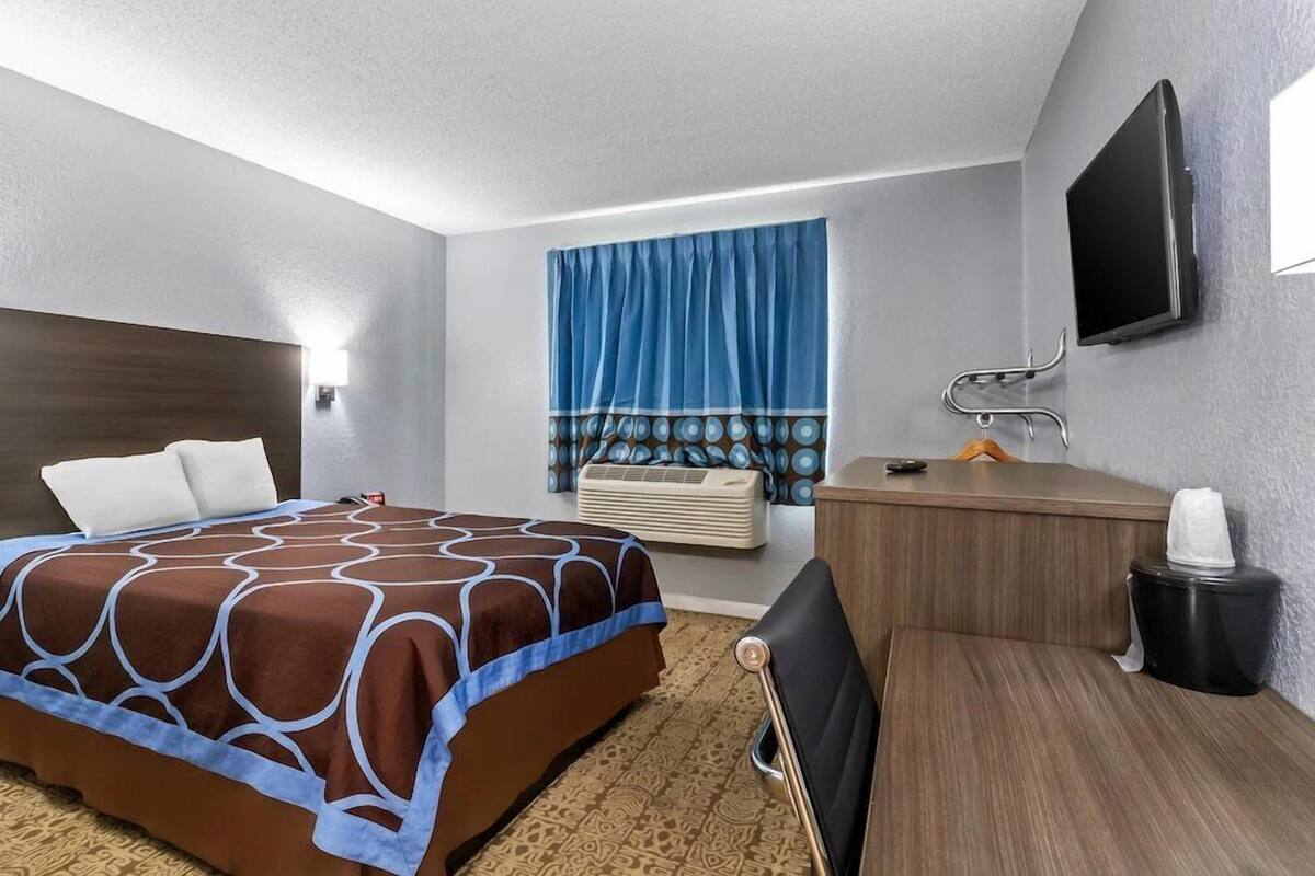 Habitación de hotel con cama, escritorio, silla, televisor, ventana con cortinas y un cesto de basura.