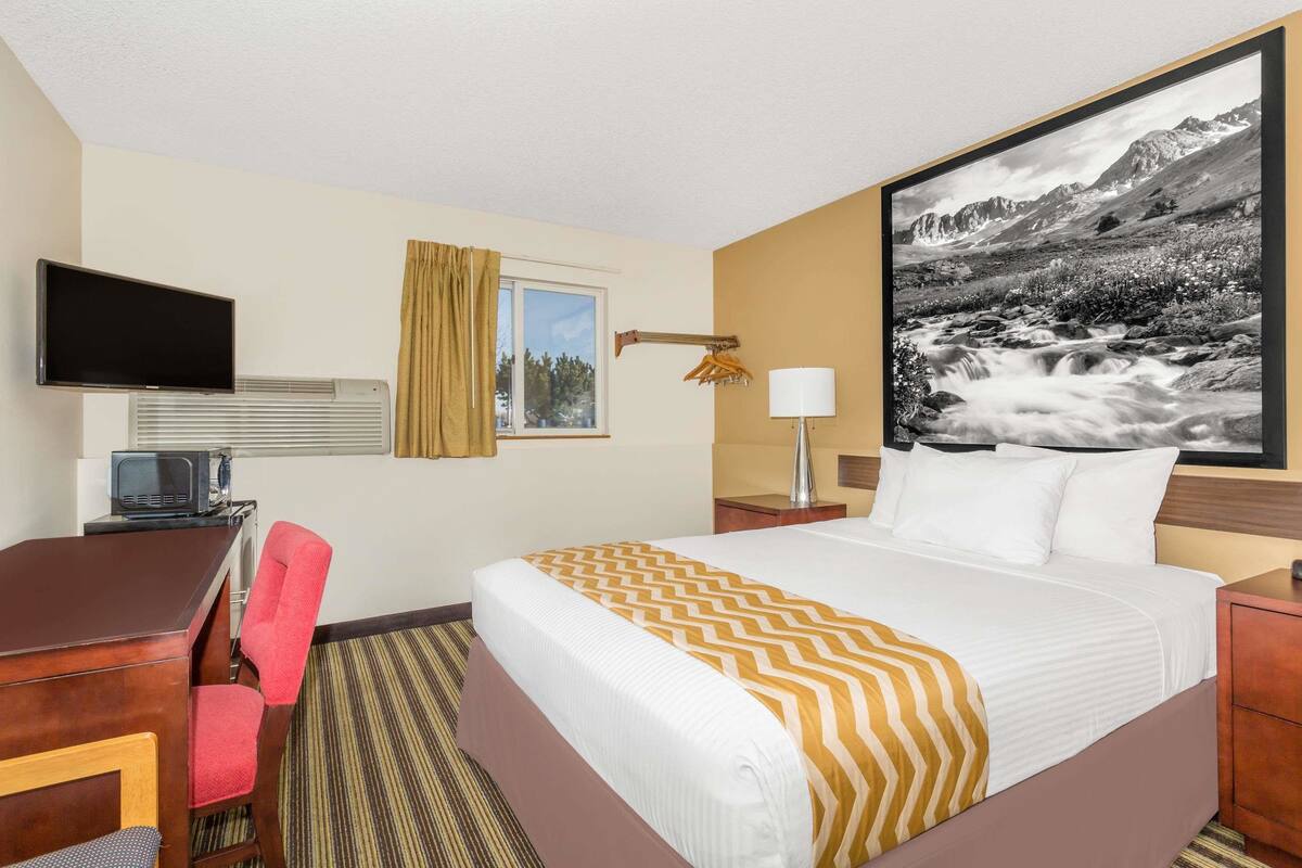 Une chambre d’hôtel comprenant un lit, un bureau, une chaise, une télévision, une lampe et un tableau représentant un paysage de montagne.