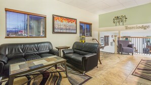 A spacious living area with a leather sofa, a glass coffee table, and a rug. There are two windows with blinds, a painting on the wall, and a Travelodge sign above the entrance. - Travelodge by Wyndham Loveland/Fort Collins Area (Loveland)