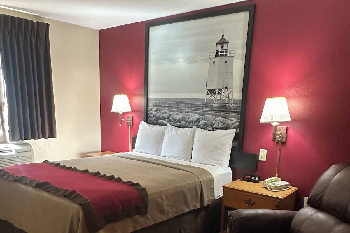 Camera d'albergo con un letto, due lampade sui comodini, un tavolino, una poltrona in pelle e un grande specchio che riflette un faro.