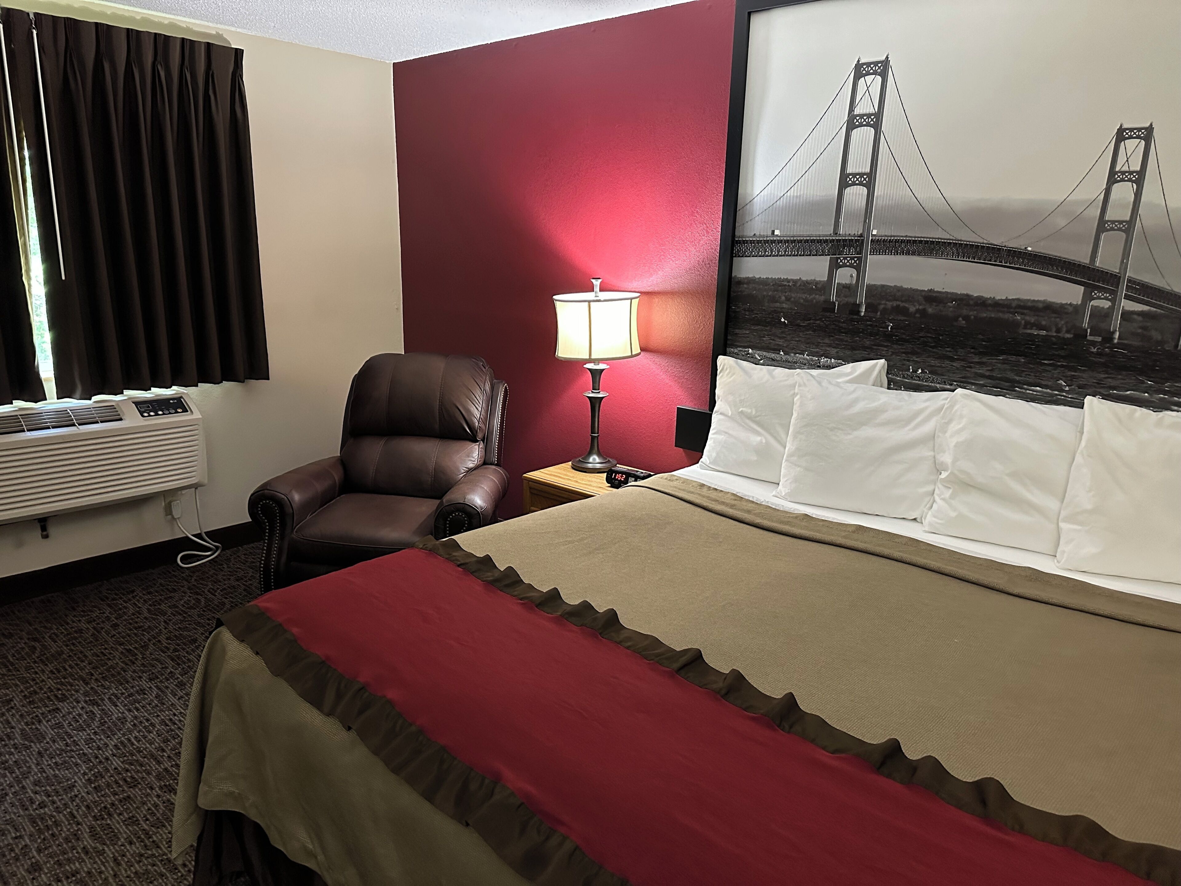 A hotel room with a bed, a chair, a desk with a lamp, a wall-mounted air conditioner, and a large framed picture of a bridge.