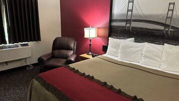 A hotel room with a bed, a chair, a desk with a lamp, a wall-mounted air conditioner, and a large framed picture of a bridge.