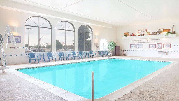 An indoor swimming pool with clear blue water, surrounded by blue chairs and tables, large arched windows, and a white tiled wall with decorative items.