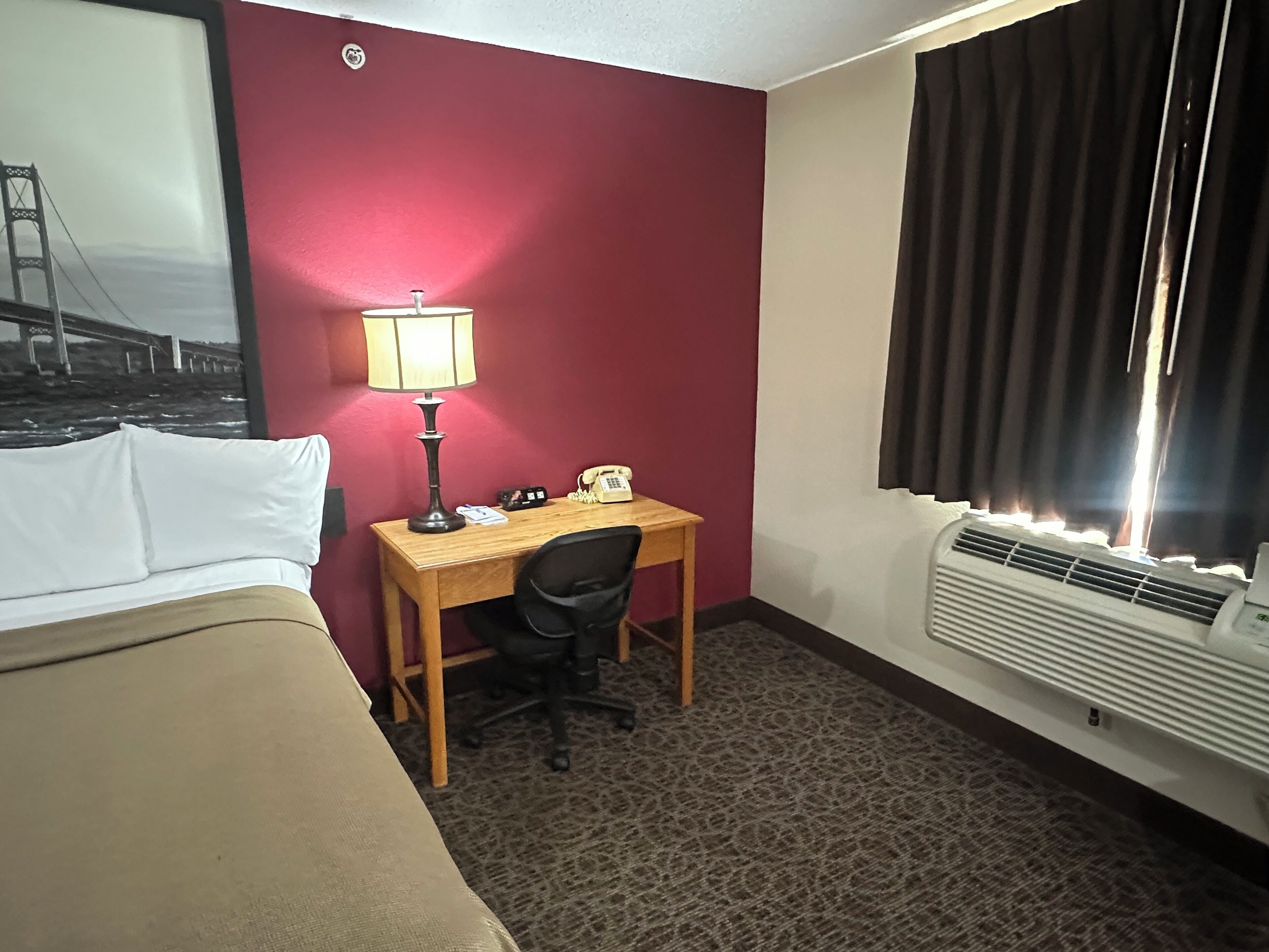 Een hotelkamer met een bed, een bureau met lamp, een stoel, een telefoon en een raam met gordijnen.
