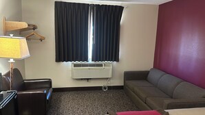 A hotel room with a gray sofa, a brown leather chair, a desk with a lamp, a window with dark curtains, and an air conditioning unit.