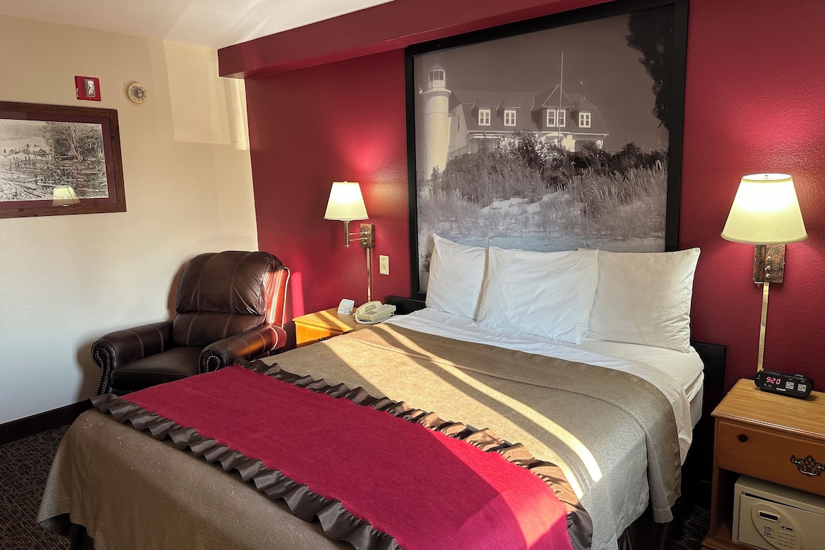 Camera d'albergo con un letto, una poltrona, un comodino con una lampada e un quadro appeso al muro.