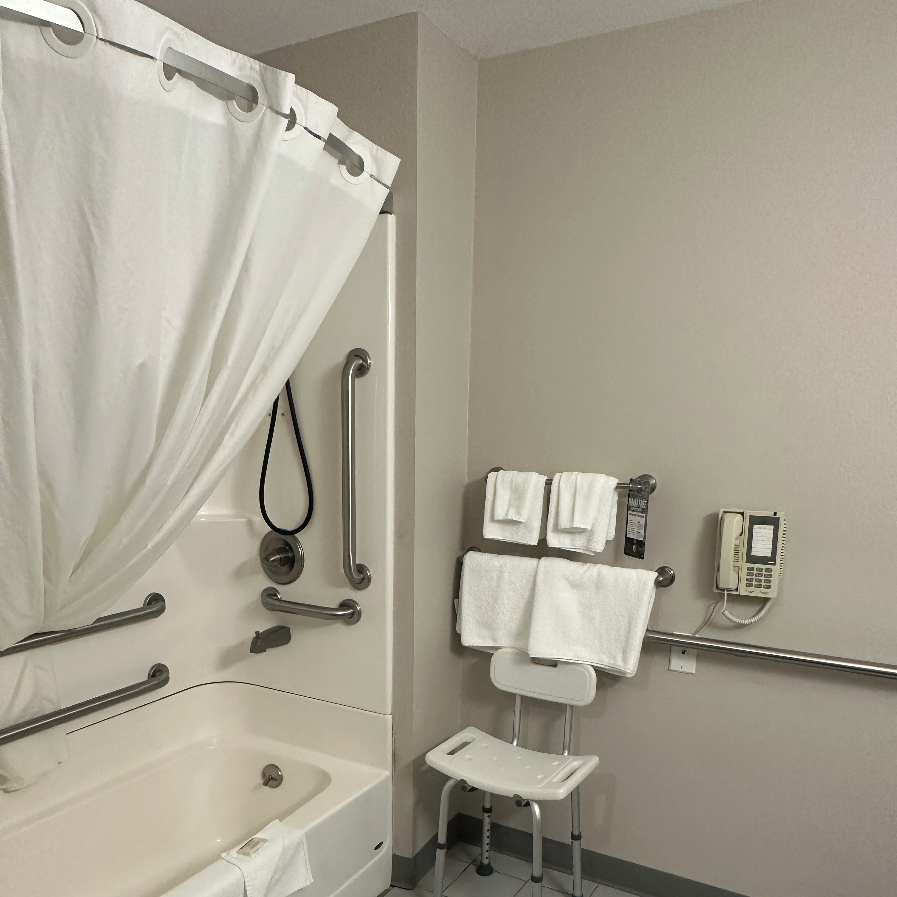Een badkamer met een bad, douchegordijn, handgrepen, een leuning, een toilet met een verhoogd zitje, een klein opstapje en een telefoon.