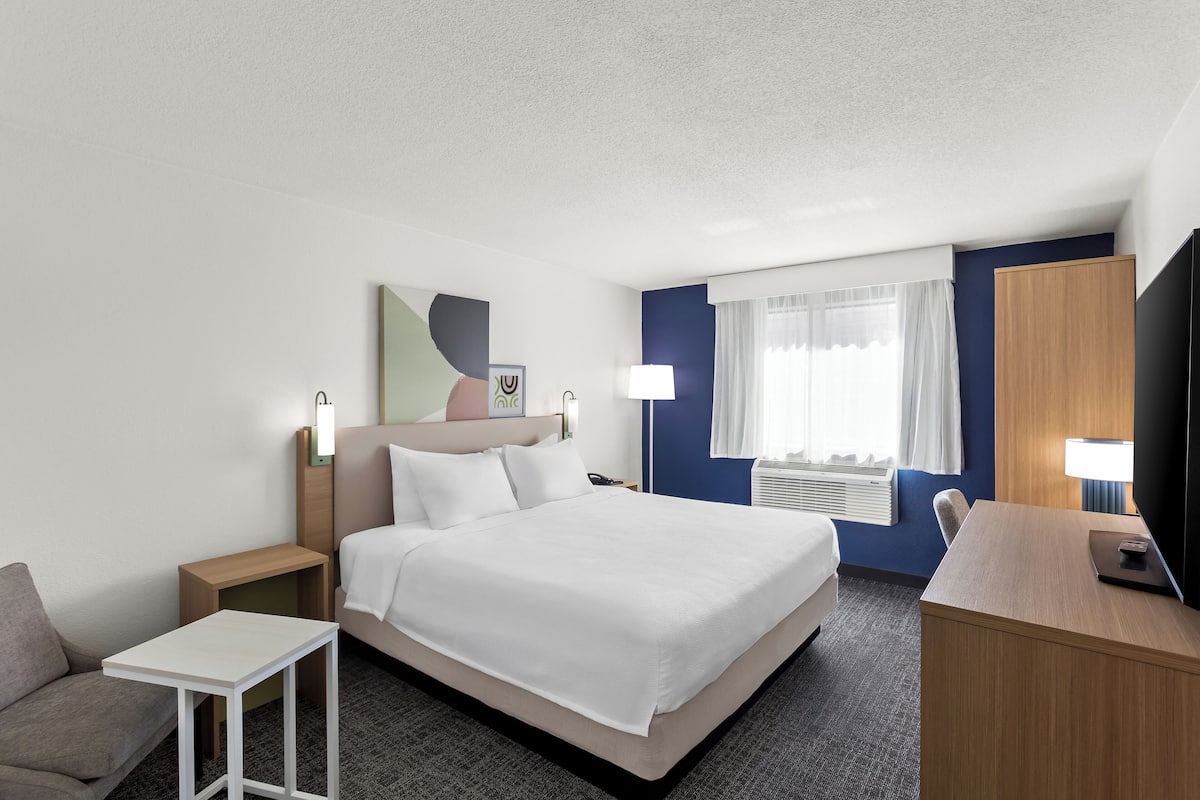 A hotel room with a bed, a desk, a chair, a small table, a TV, and a window with curtains.