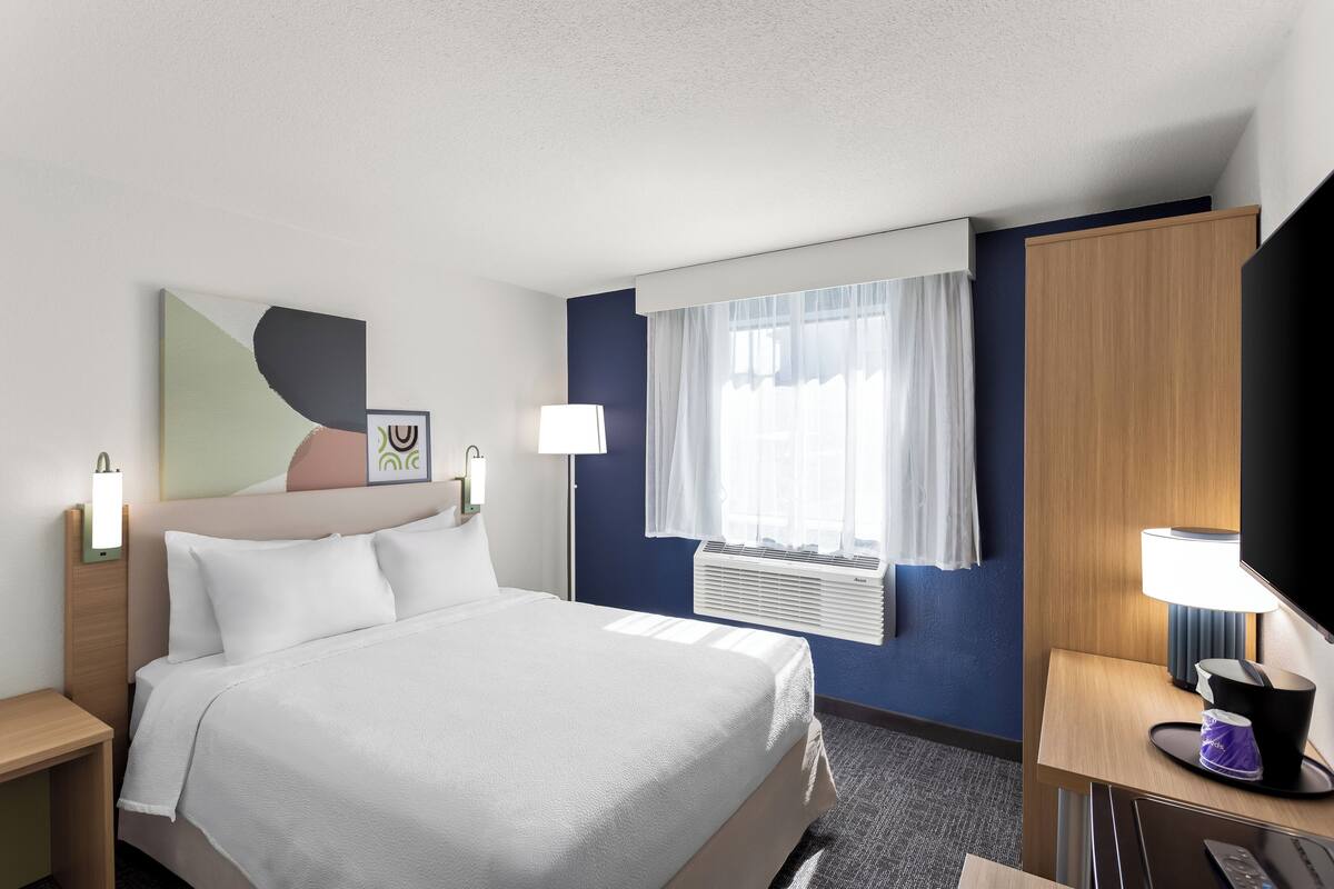 A hotel room with a bed, bedside tables, a desk with a lamp, a flat-screen TV, a window with curtains, and a wall-mounted air conditioner.
