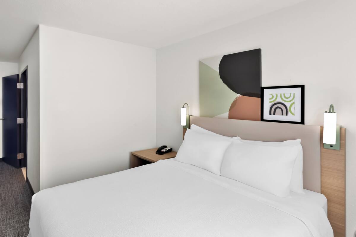 A hotel room with a bed, bedside table, phone, wall art, and a door.