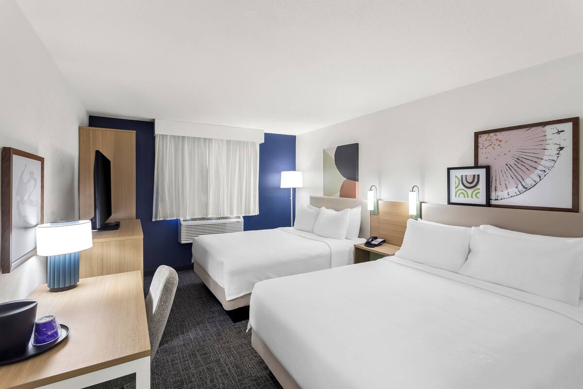 A hotel room with two beds, a desk with a lamp, a chair, a TV, and a window with curtains.
