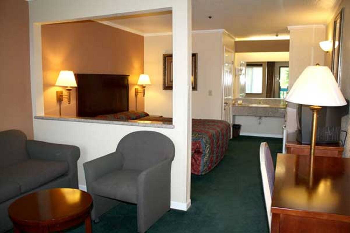 A hotel room with a sofa, armchair, and a small table. There is a bed with a colorful blanket and a mirror above it.