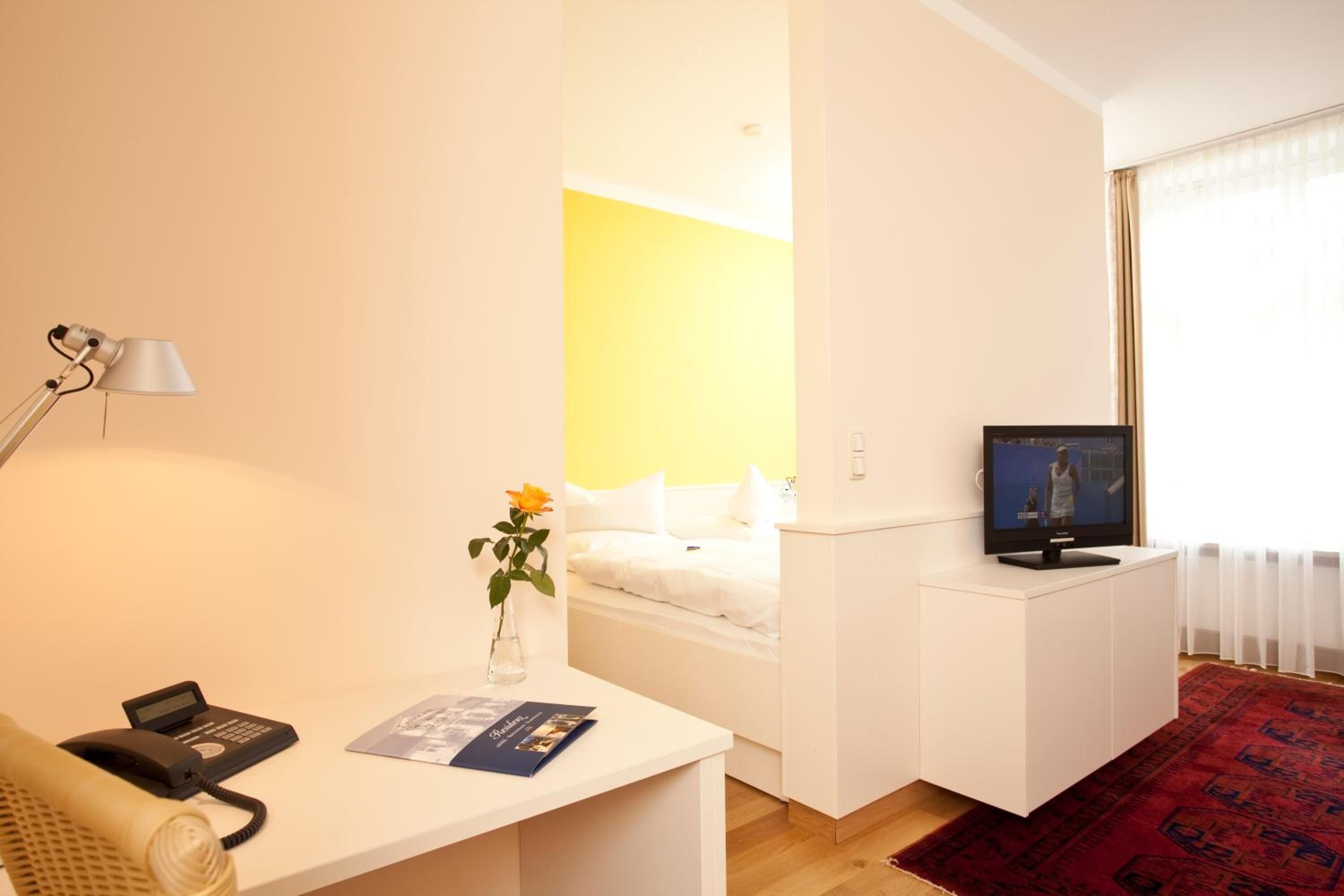 Ett hotellrum med en säng, ett skrivbord med en lampa, en tv och ett litet bord med en vas med blommor.