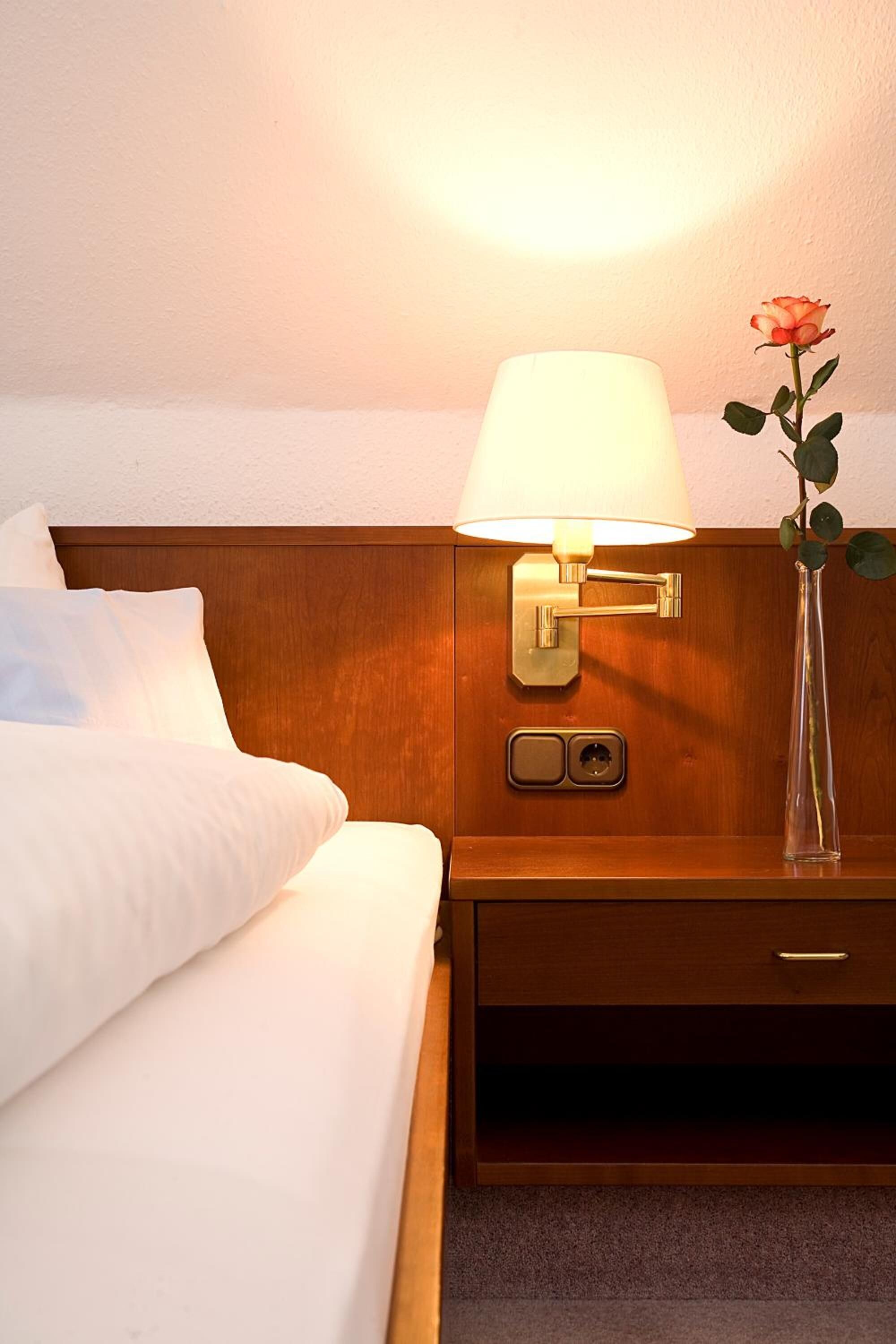 A neatly made bed with white linens, a bedside lamp with a white shade, a vase with a single rose, and a wooden nightstand with a drawer.