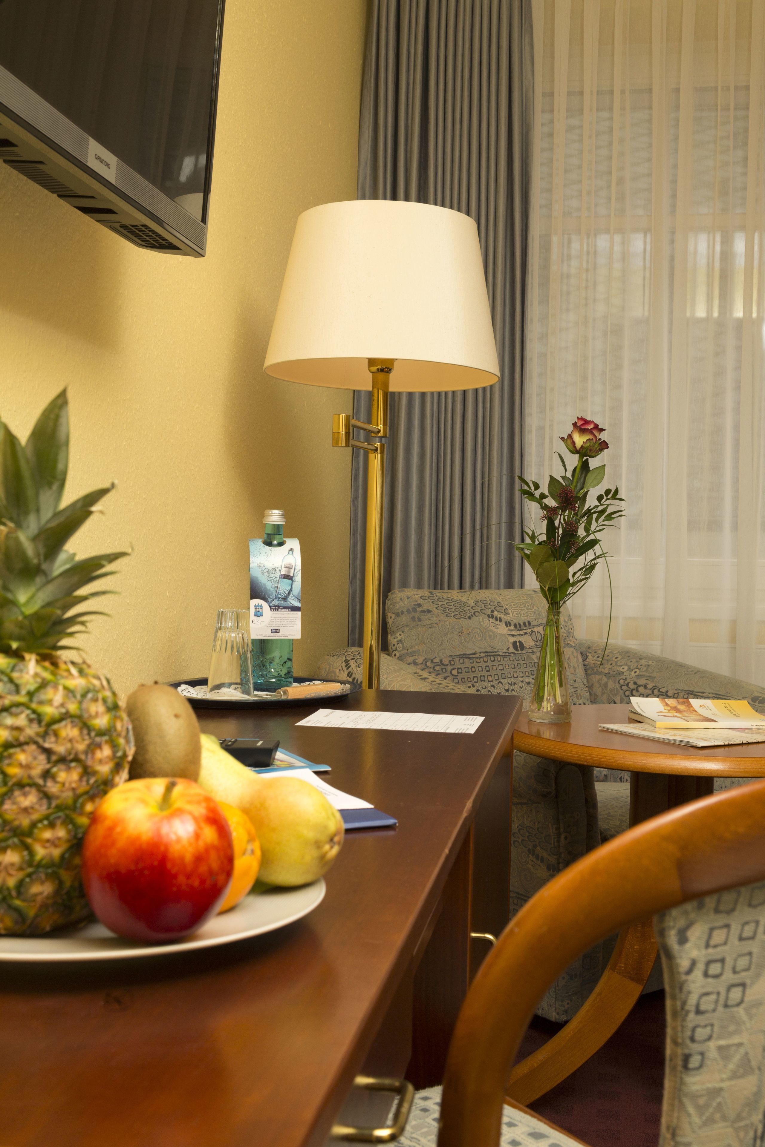 A hotel room with a dining table, a lamp, a vase with a flower, and a fruit plate.