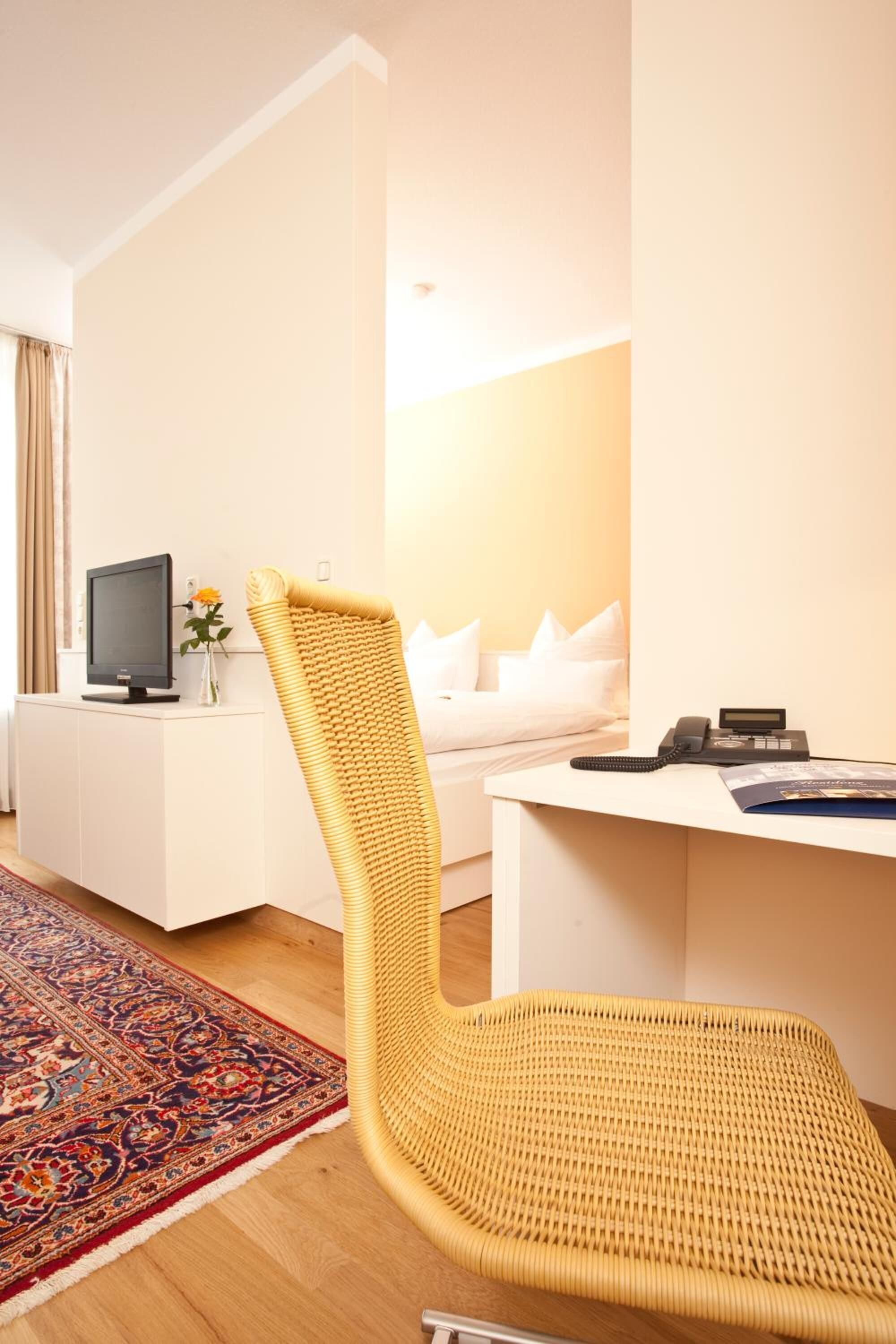 A hotel room with a yellow woven chair, a white desk, a television, and a bed with white bedding.