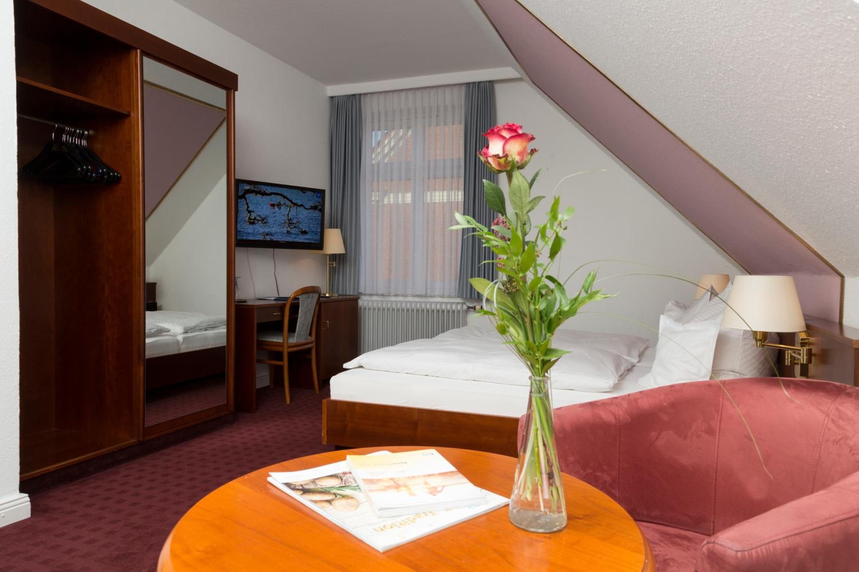 A hotel room with a bed, a desk, a chair, a round table with a vase of flowers, and a large mirror.