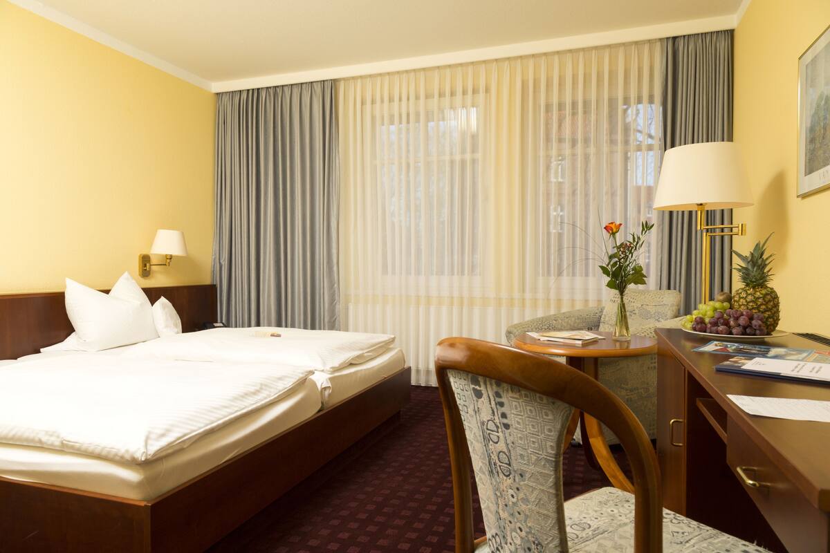 A hotel room with a bed, a desk with a lamp, a chair, and a table with fruits.