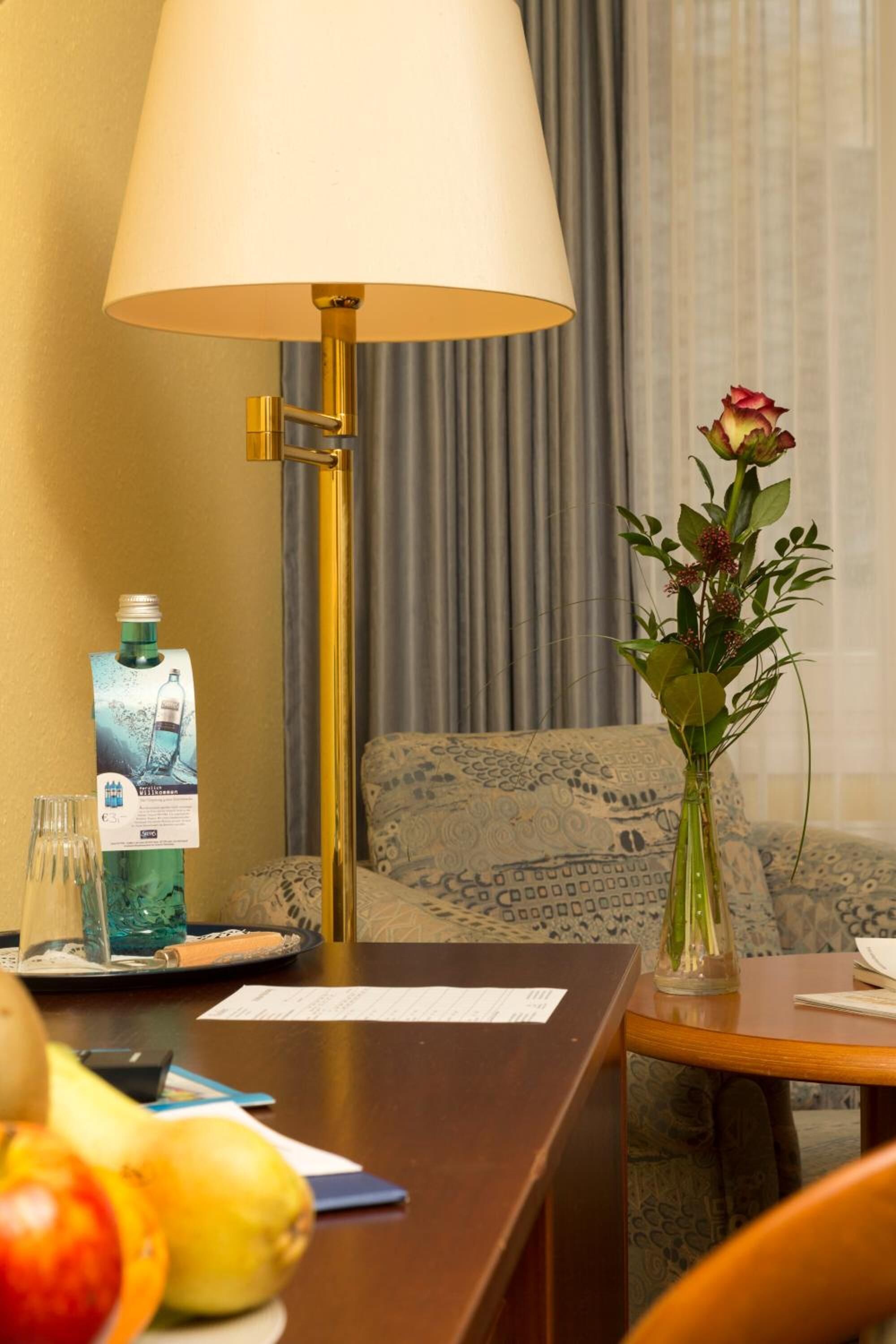 A hotel room with a wooden desk, a lamp, a vase with flowers, a bottle of water, and a patterned sofa.