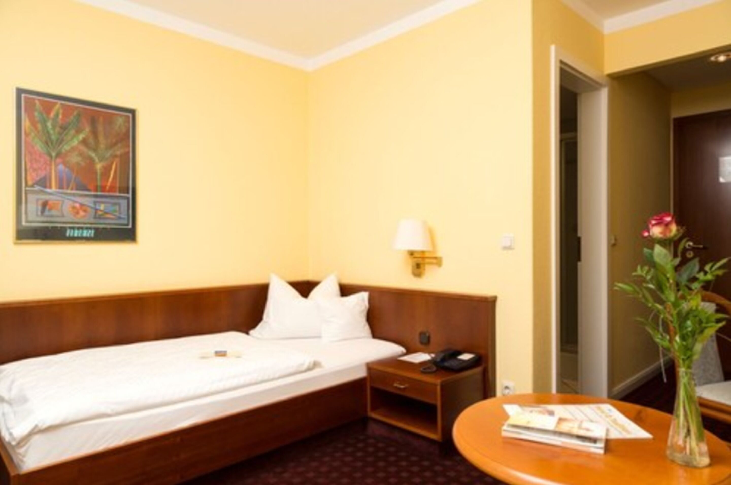 A hotel room with a single bed, a bedside table with a lamp, a small table with a vase and a book, and a painting on the wall.