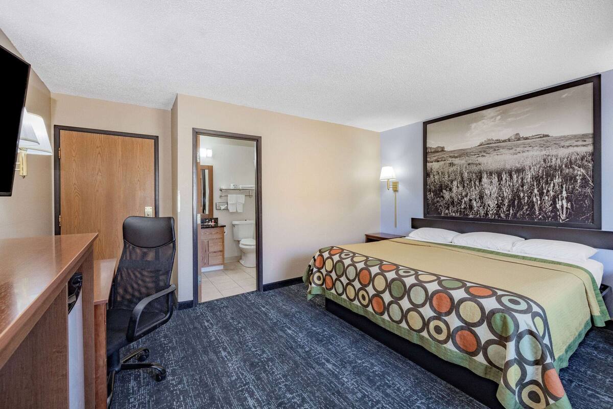 A hotel room with a desk, office chair, bed with patterned bedding, and a large landscape painting on the wall.
