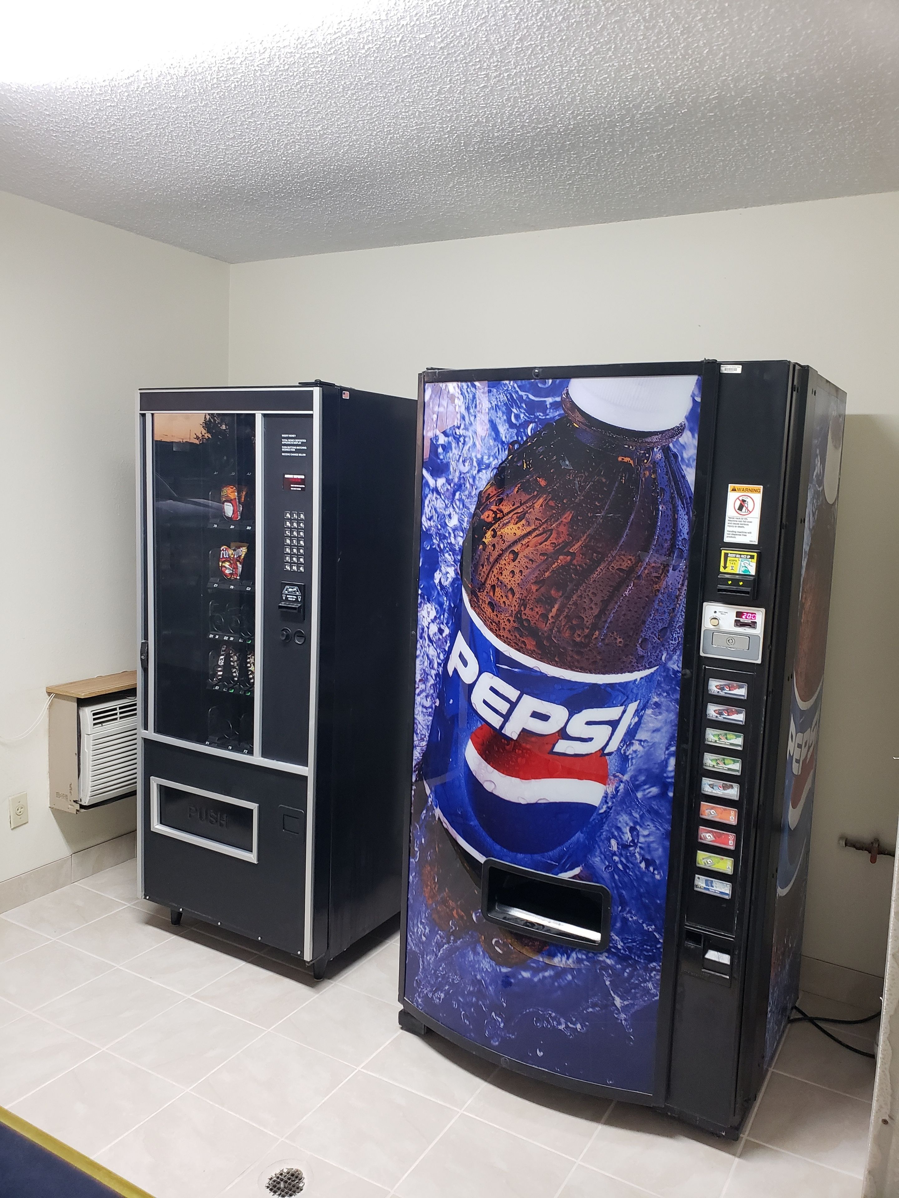 Two vending machines, one with a Pepsi logo and the other with a different design, in a room with tiled flooring and a white ceiling.