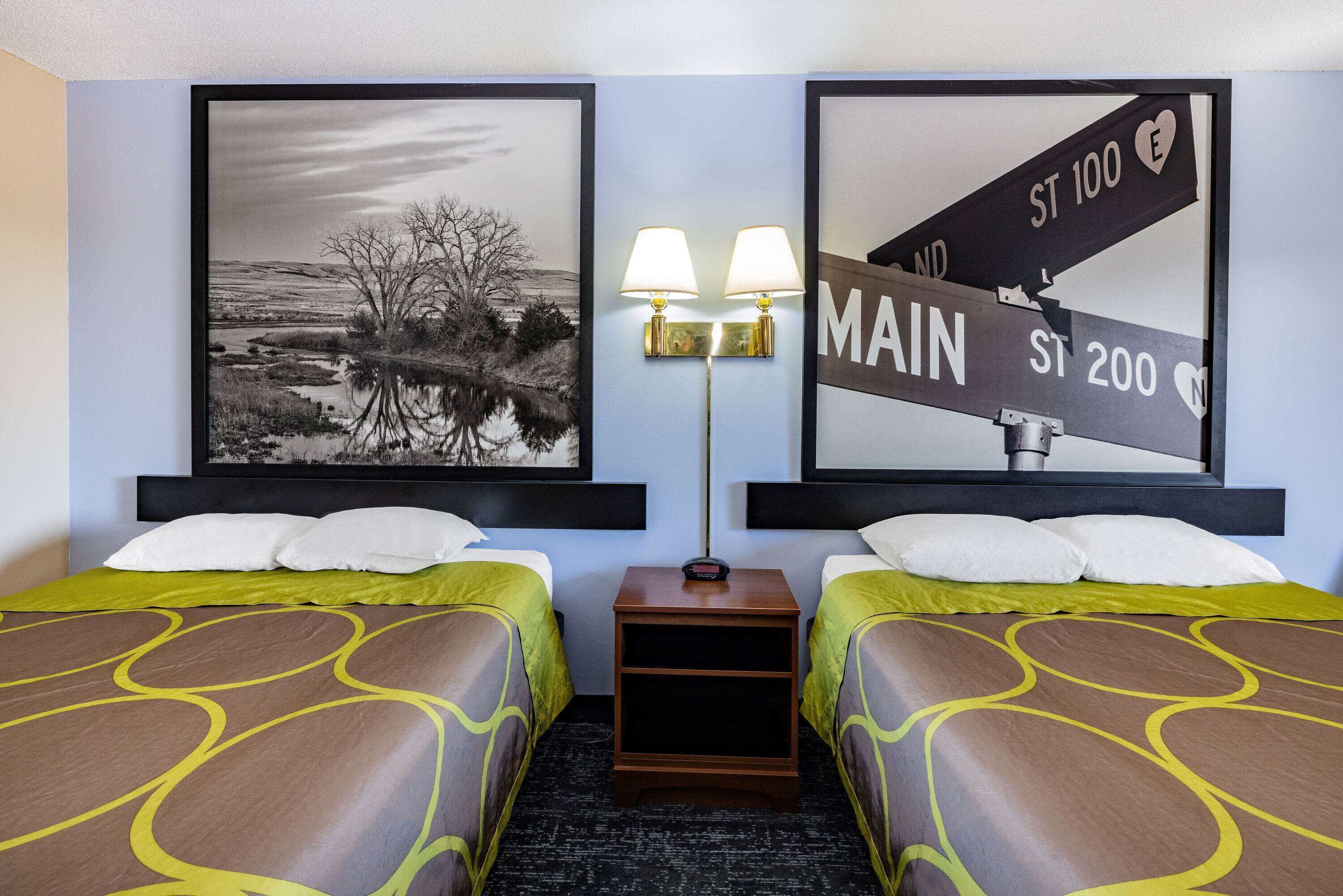 A hotel room with two beds, a nightstand, and wall art depicting street signs.