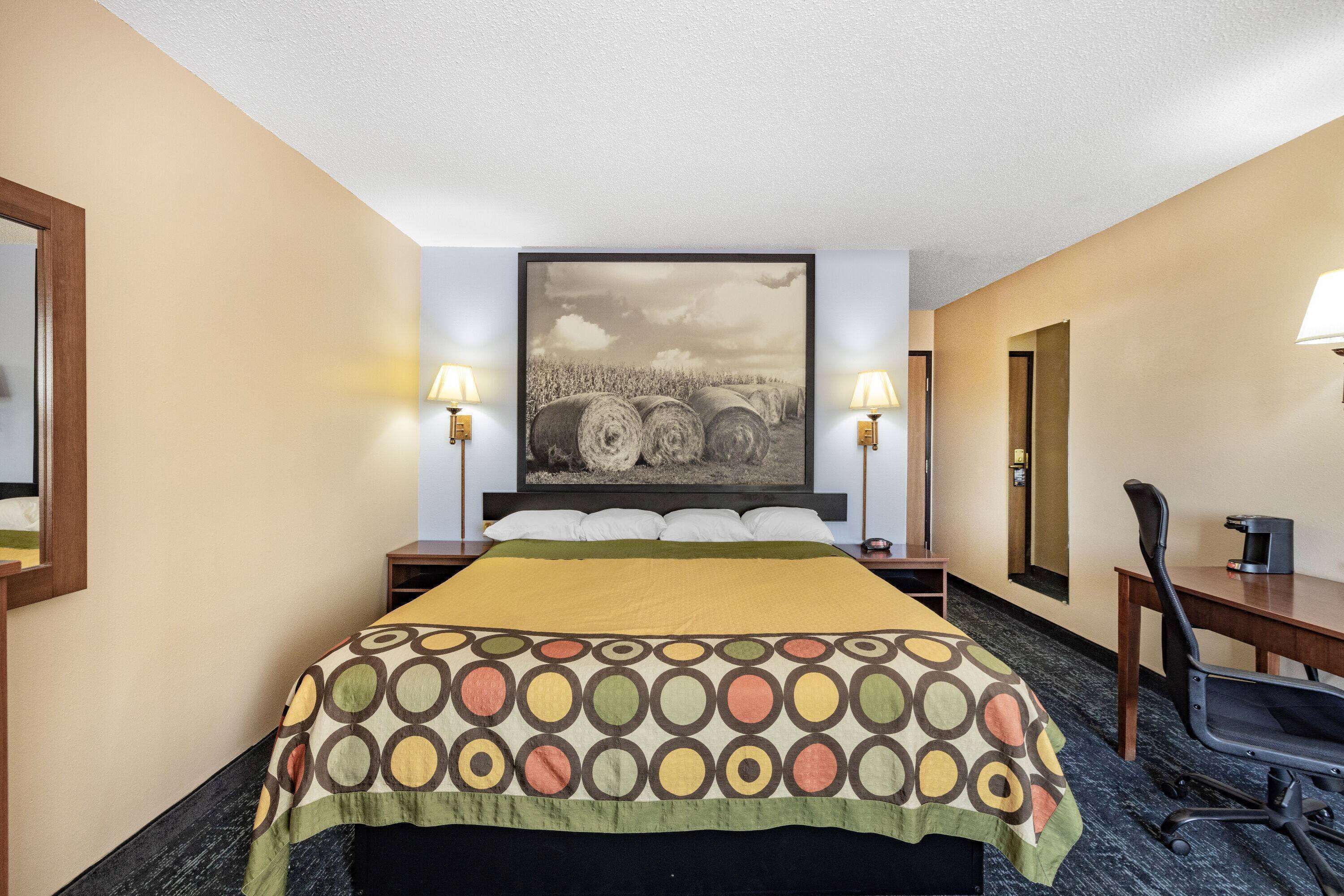 A hotel room with a large bed, two bedside lamps, a desk with a chair, and a large framed picture on the wall.