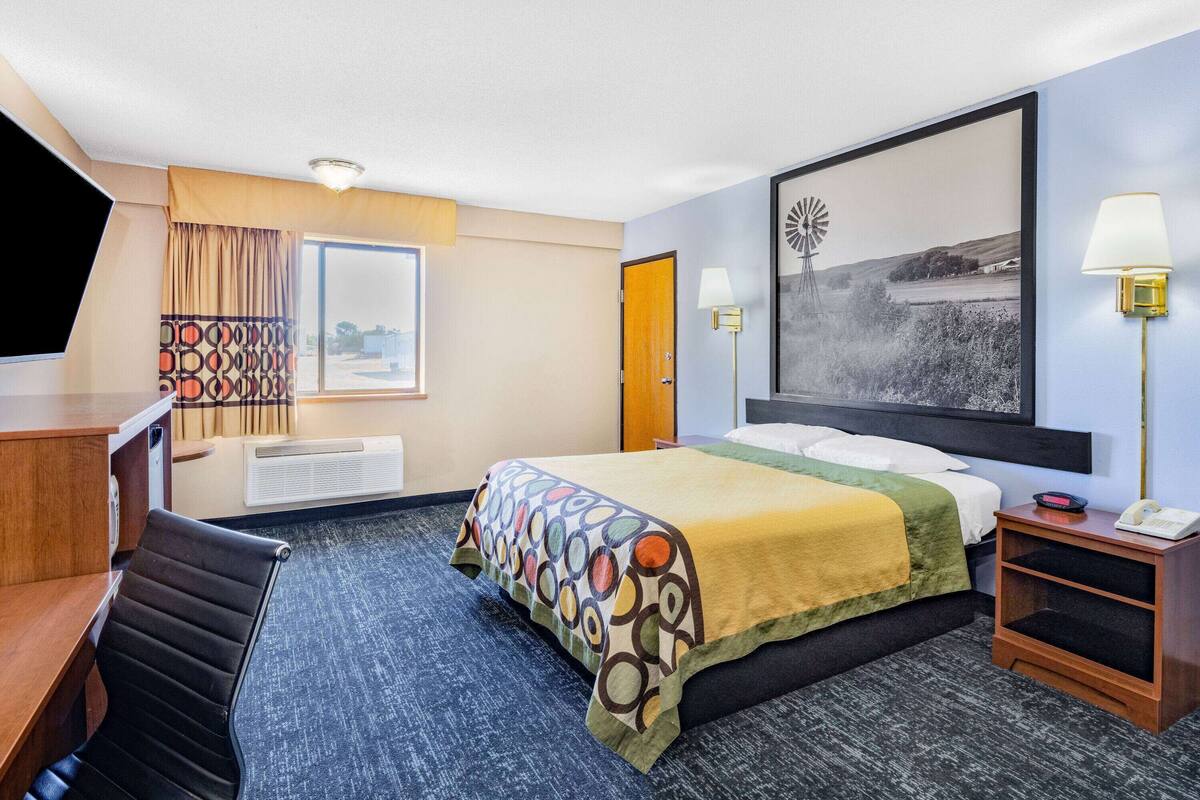 A hotel room with a bed, a desk, a chair, a desk lamp, a wall-mounted lamp, and a large framed picture.
