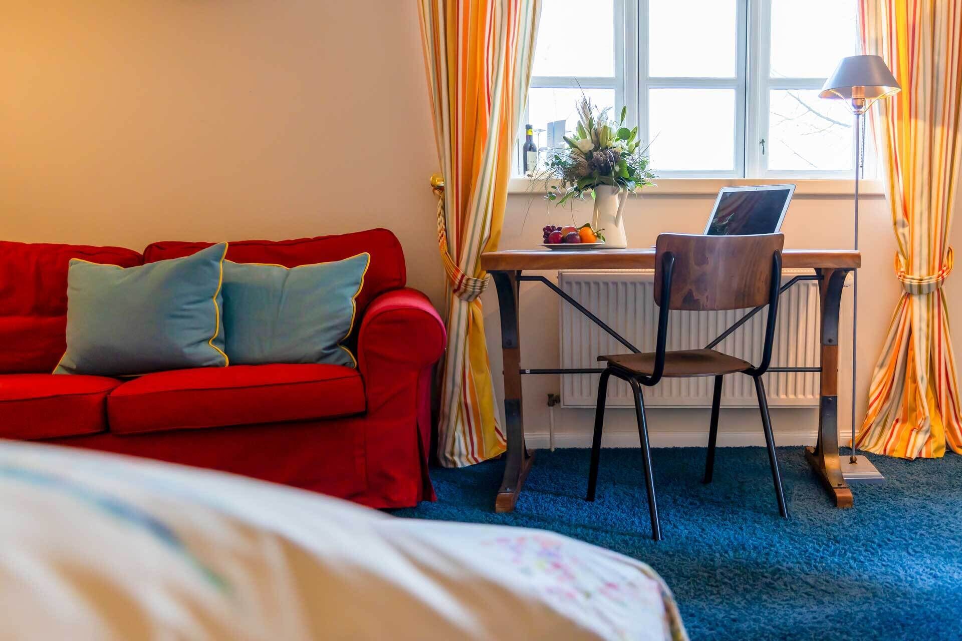A room with a red sofa, a wooden desk with a chair, a laptop, a vase with flowers, and a window with curtains.