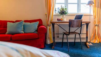 A room with a red sofa, a wooden desk with a chair, a laptop, a vase with flowers, and a window with curtains.