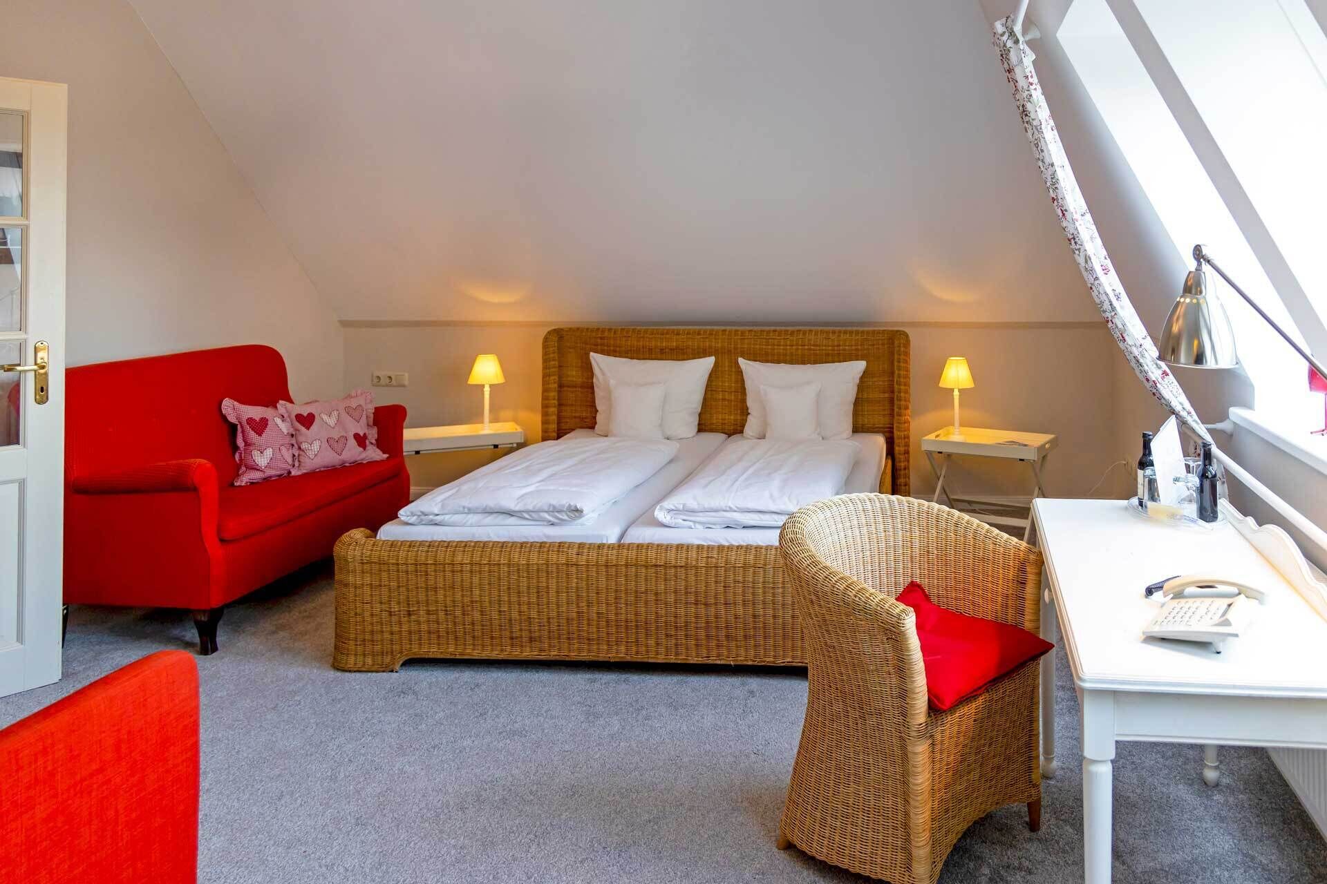 Un dormitorio con una cama, dos mesitas de noche, un sofá rojo, un sillón de mimbre y un escritorio con una lámpara.