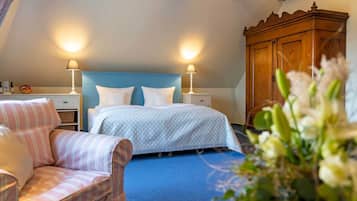 Una habitación de hotel con una cama, dos lámparas de noche, un armario de madera, un sillón a rayas y una mesita con un jarrón de flores.