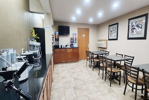 A hotel lobby with a reception desk, a coffee machine, a refrigerator, a dining area with tables and chairs, and a television mounted on the wall.