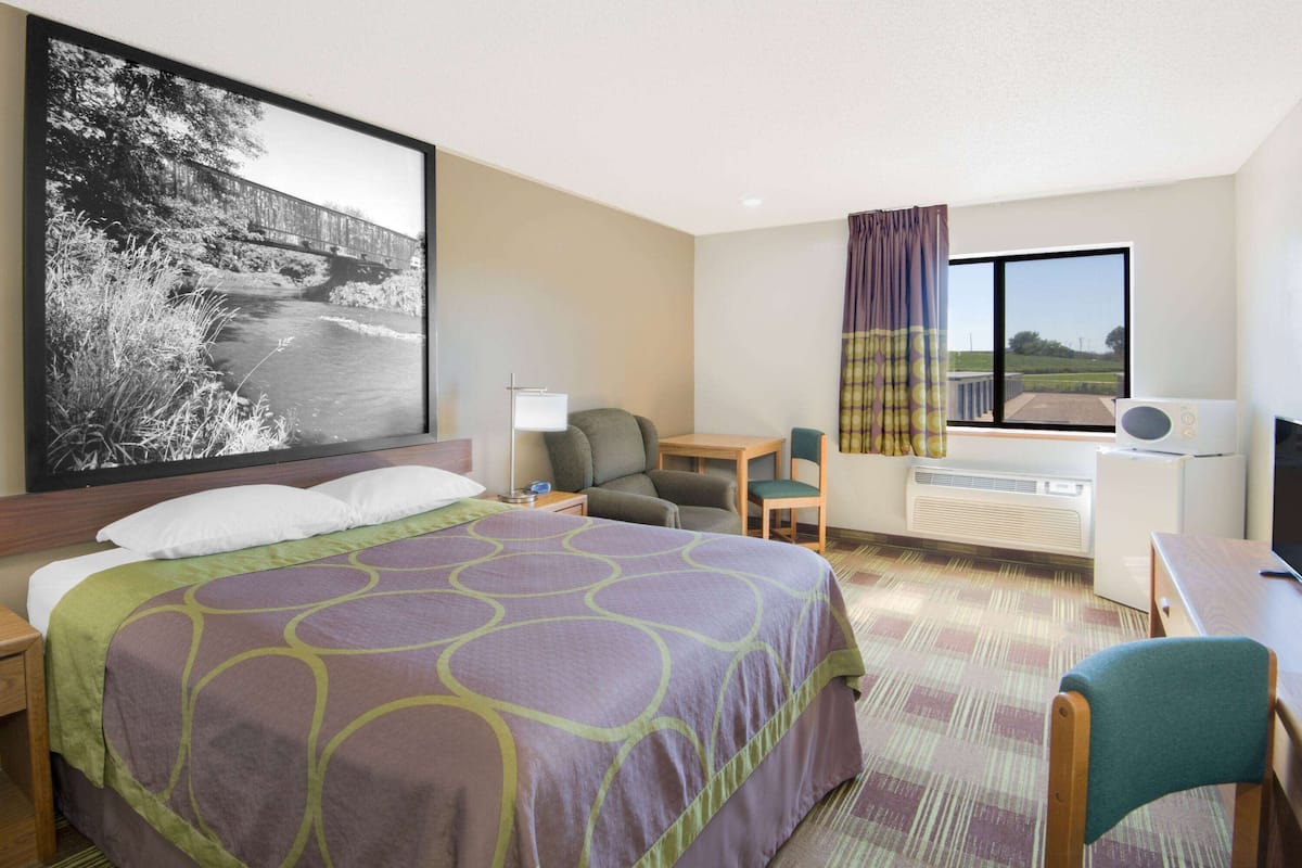 Quarto de hotel com uma cama grande, uma escrivaninha, uma cadeira, televisão e janela com vista para uma ponte e vegetação.