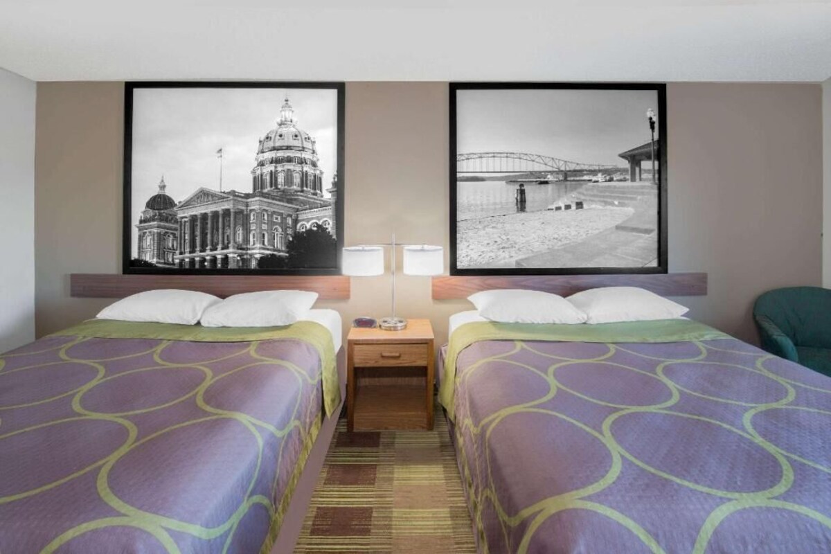 Une chambre d’hôtel avec deux lits, une table de chevet, deux lampes fixées au mur et deux tableaux encadrés accrochés au mur.