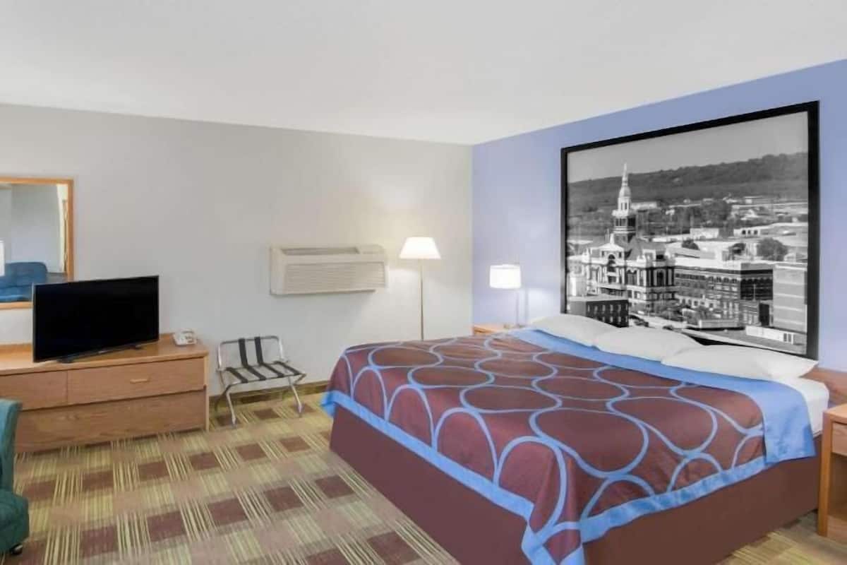 Quarto de hotel com cama, televisão sobre uma cômoda de madeira, cadeira, escrivaninha e um quadro grande na parede.