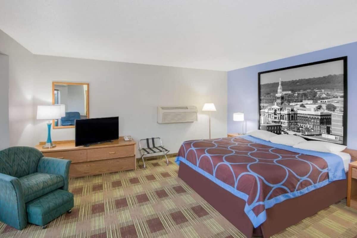 Quarto de hotel com uma cama, televisão em uma cômoda, uma cadeira, escrivaninha e uma grande moldura na parede.