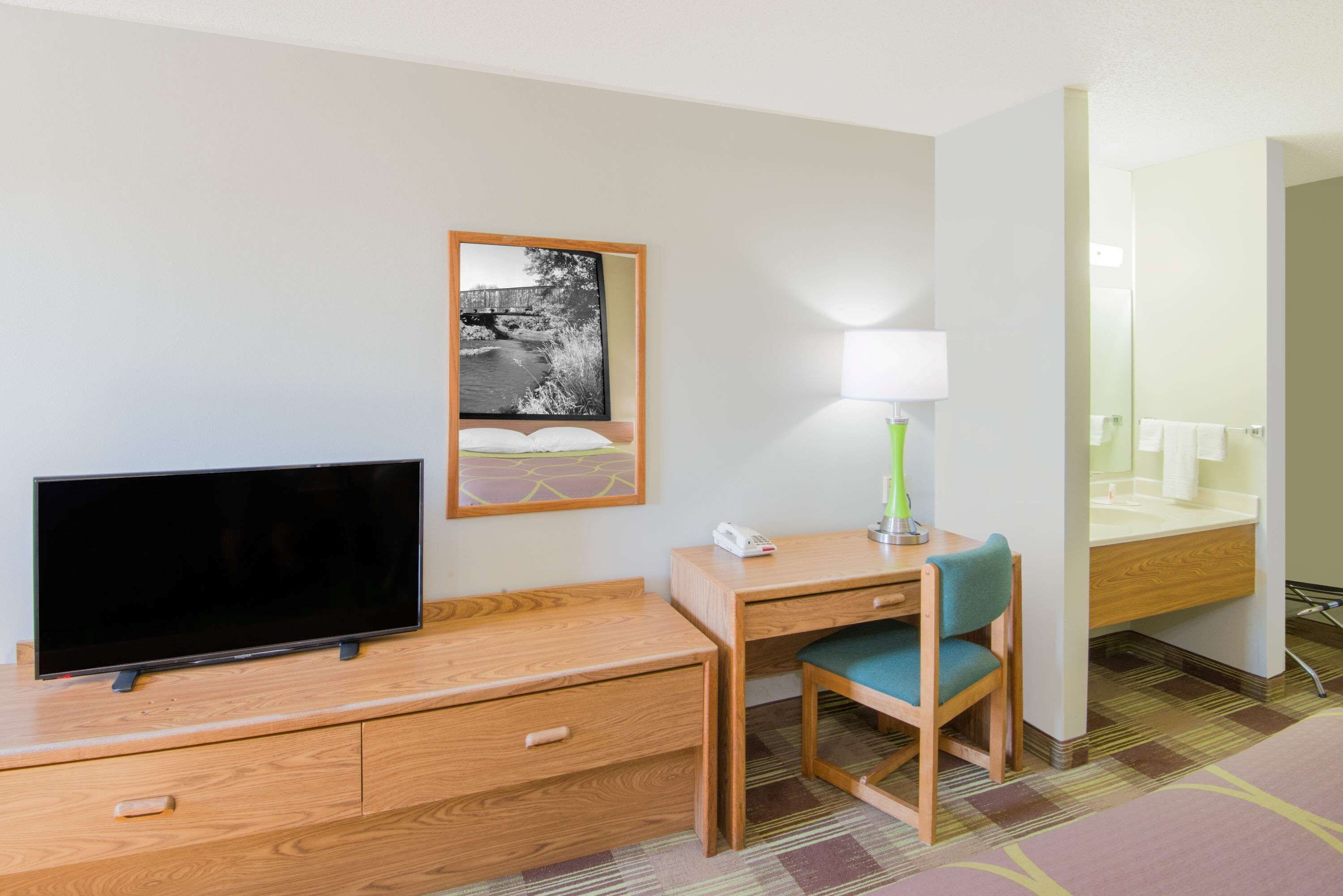 A hotel room with a flat-screen TV, a desk with a chair, a lamp, a painting, and a bathroom with a mirror and sink.