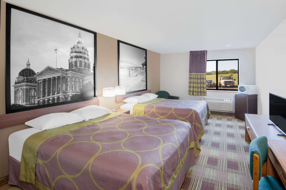 Quarto de hotel com duas camas, uma escrivaninha, uma cadeira, televisão e uma janela grande com vista para um edifício e uma ponte.