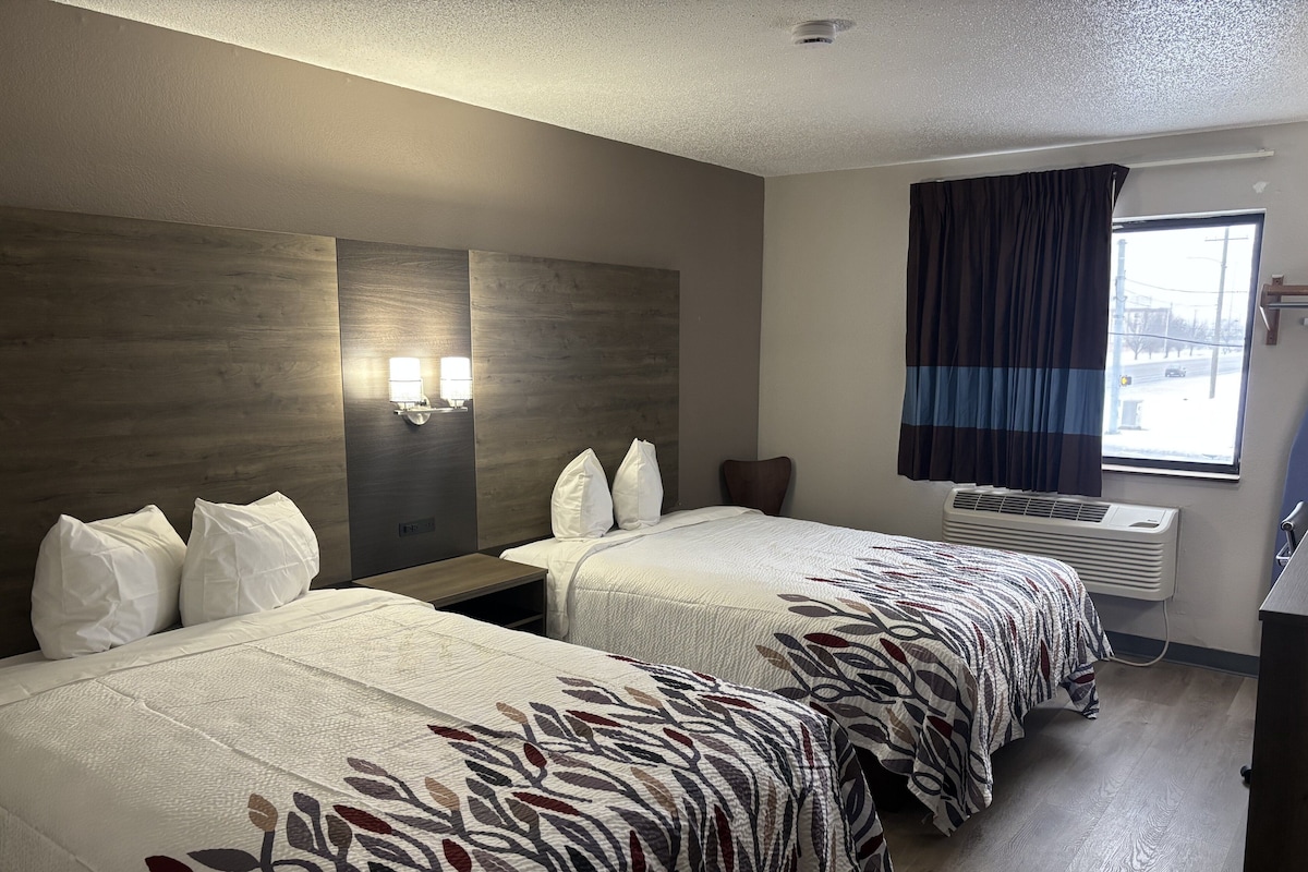 Ein Hotelzimmer mit zwei Betten, einem Nachttisch, einer Wandlampe, einem Fenster mit Vorhängen und einer Klimaanlage.