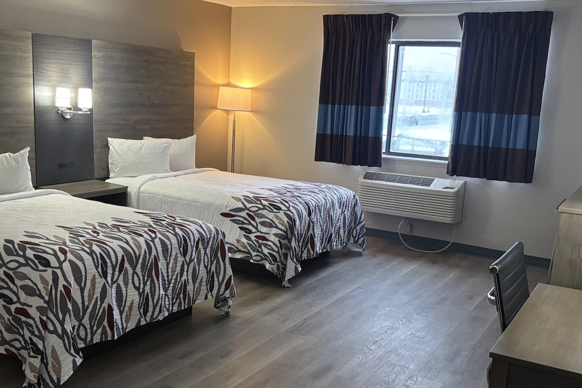 Ein Hotelzimmer mit zwei Betten, einem Nachttisch, einer Lampe, einer Klimaanlage und einem Fenster mit Vorhängen.