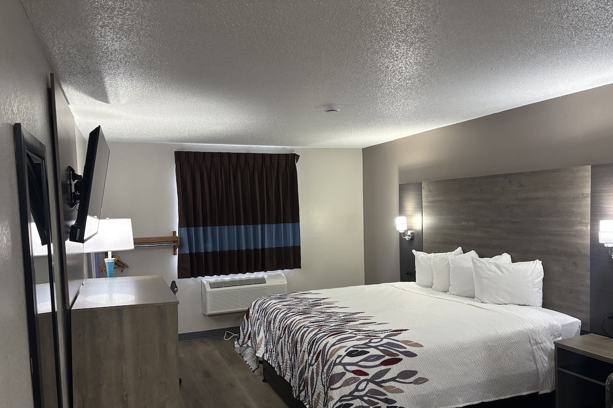 Ein Hotelzimmer mit einem großen Bett, einem Flachbildfernseher, einem Nachttisch, einer Lampe, einem Fenster mit Vorhängen und einer Wandleuchte.